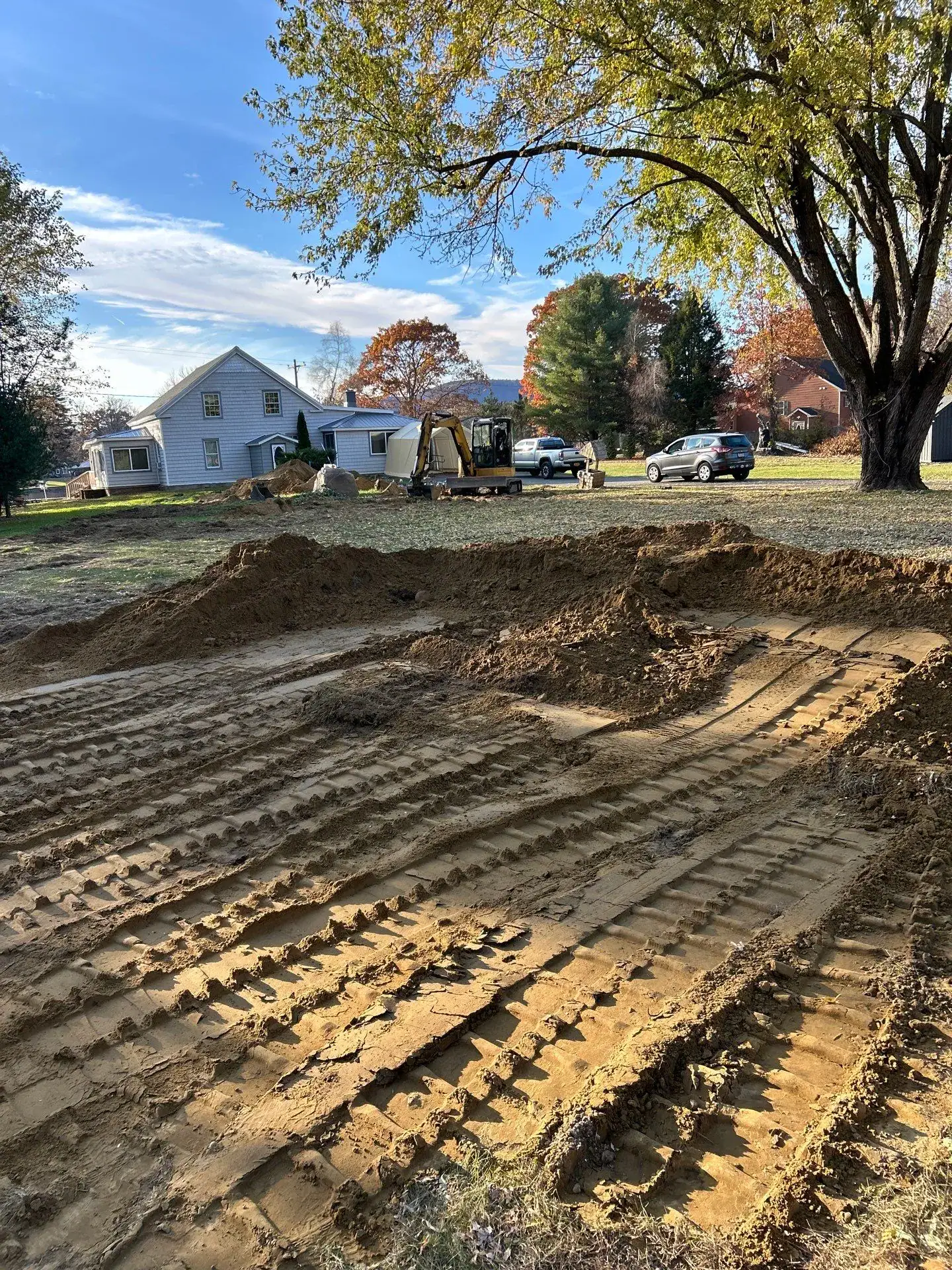 A yard with fresh dirt and deep bulldozer tracks; a small excavator, from experienced excavating contractors Saratoga County, is parked near a house in NY under a large tree with autumn leaves.
