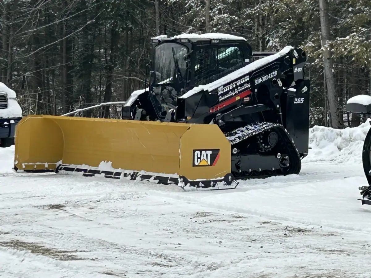 A black Caterpillar skid steer with a yellow snow plow attachment is clearing snow in a forested area. Snow covers the ground and parts of the vehicle, while NY excavating contractors Saratoga County work among the trees in the background.