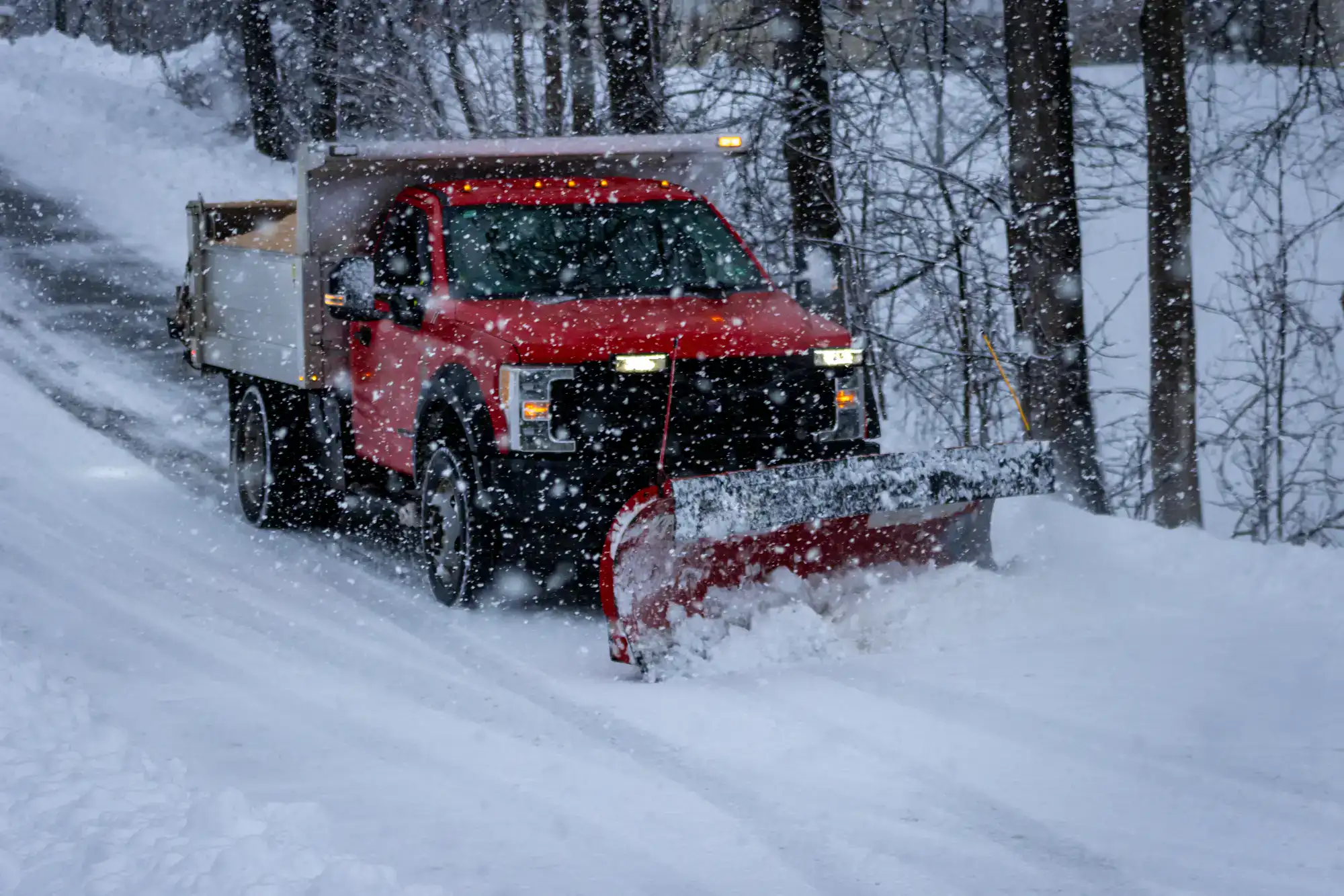 A red snowplow truck clearing snow from a tree-lined road during heavy snowfall. Snow is falling thickly, covering the ground, trees, and parts of the truck.