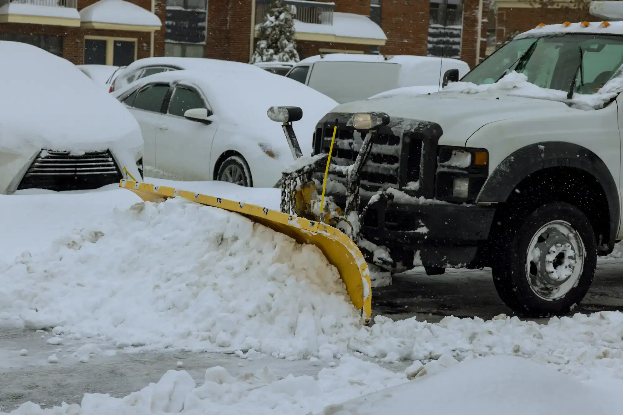 A snow plow truck clearing snow from a parking lot, with several parked cars partially covered in snow in the background on a winter day.