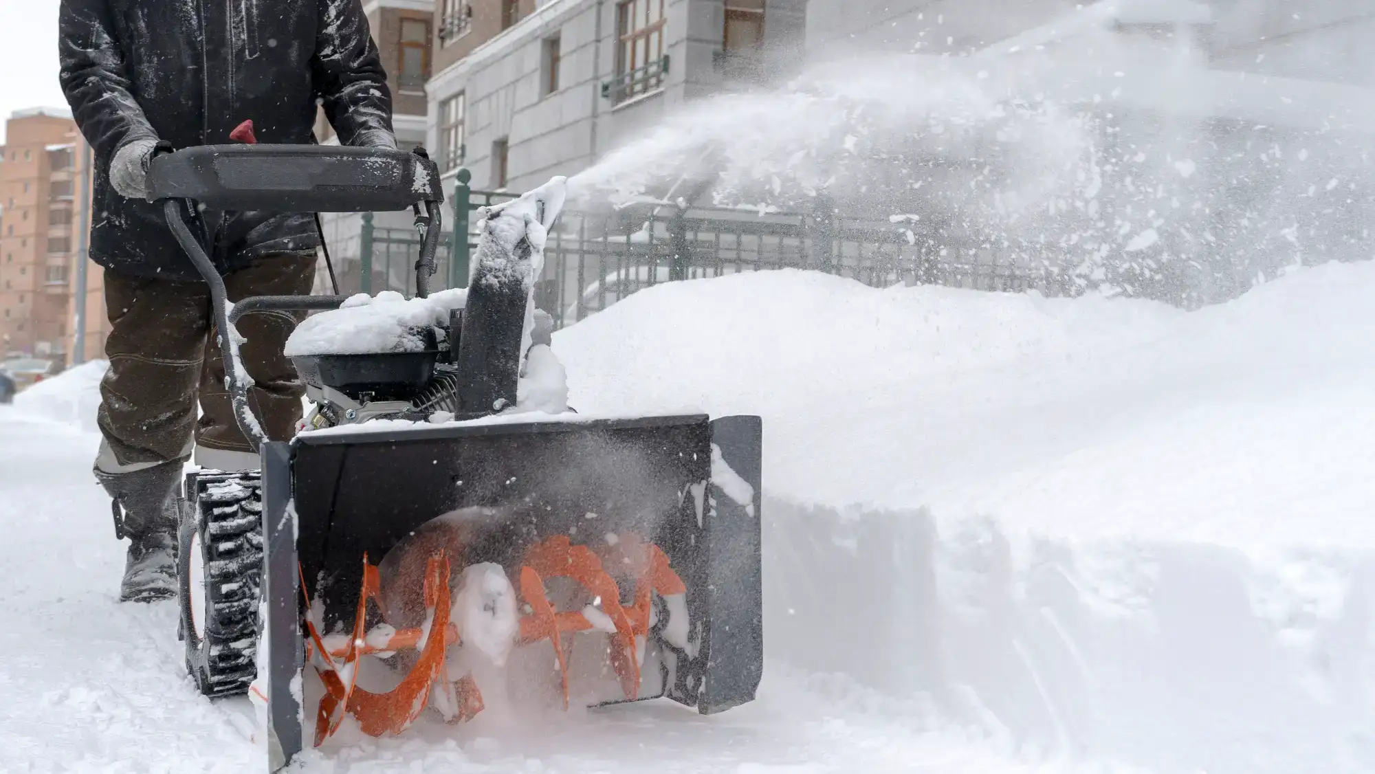 A person using a snow blower to clear a sidewalk covered in deep snow, with snow being thrown to the side near buildings in a residential area.