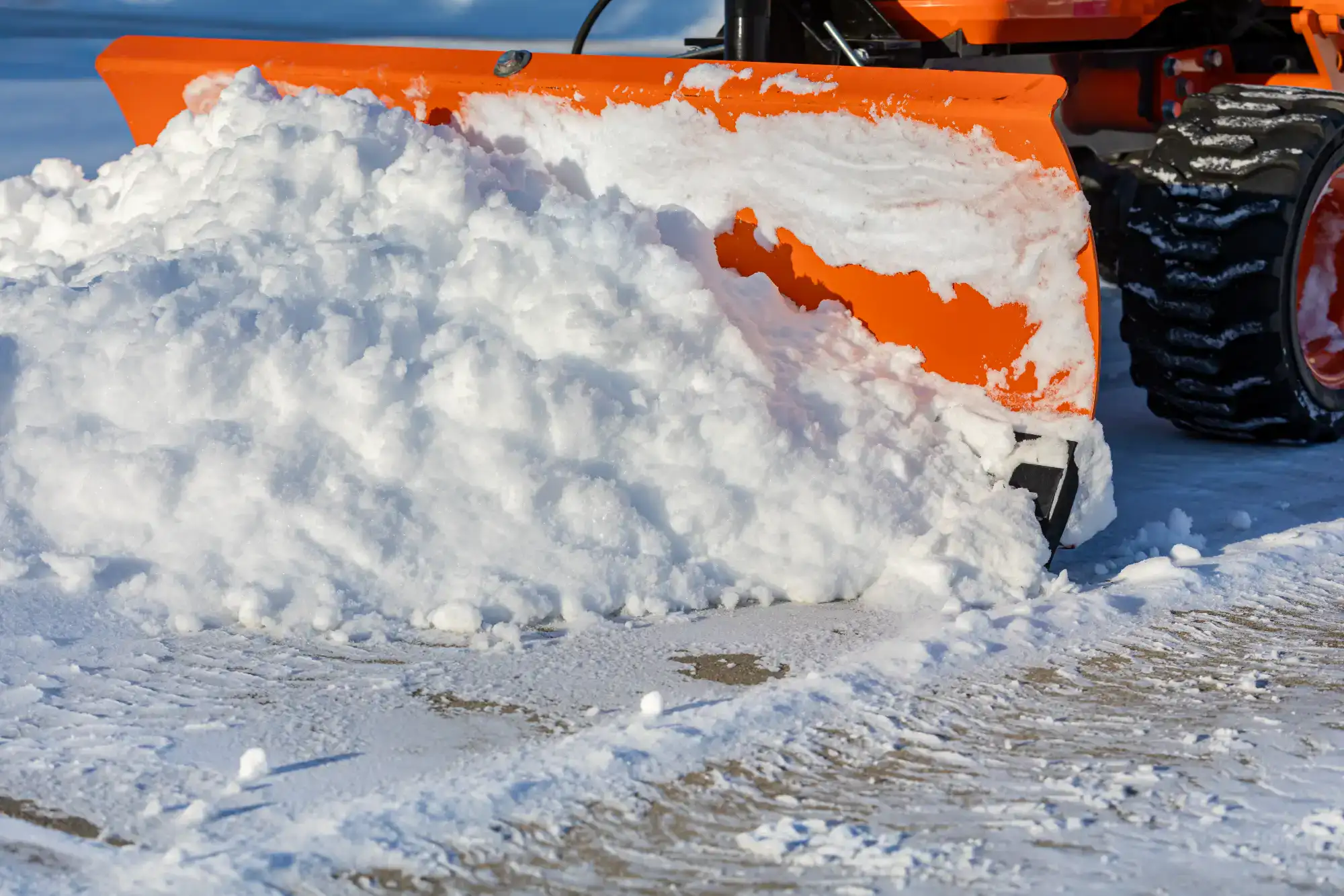 An orange snow plow pushes a large pile of snow off a paved surface on a sunny winter day.
