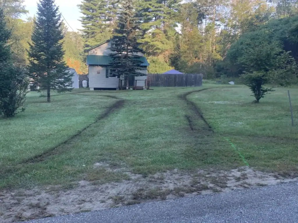 A small house with a green roof sits in a grassy yard. Curved tire tracks, possibly made by excavating contractors Saratoga County, NY, lead from the road to the house, passing several trees, with woods and a fenced area in the background.