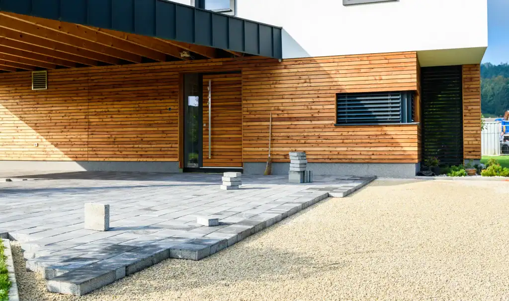 The exterior of a modern home featuring wooden slat siding and a deep carport is shown, with a partially completed grey paver driveway extending onto a bed of light gravel.